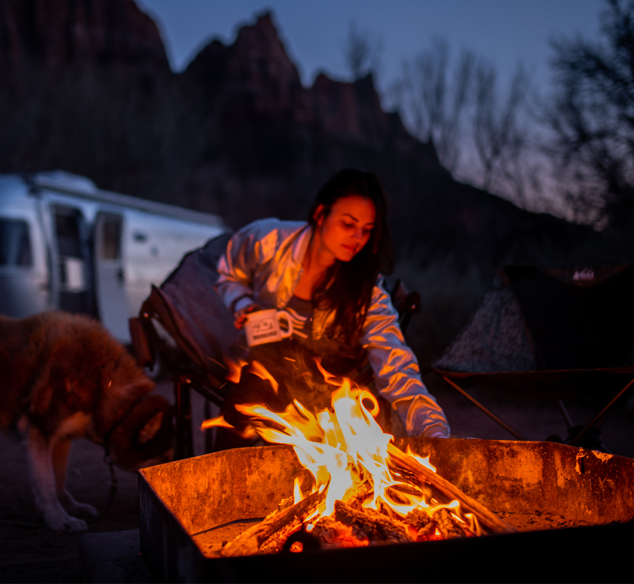 A woman sitting by a campfire with her dog