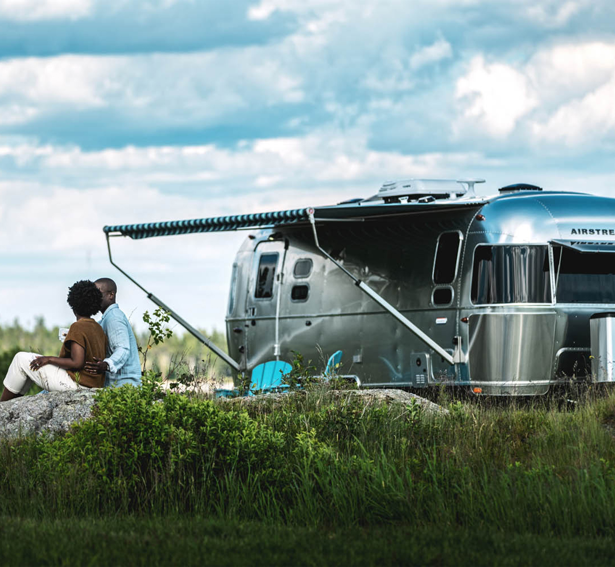 A couple sitting on a rock in the grass outside of their Airstream Travel Trailer in a grassy field with the Airstream's awning out.