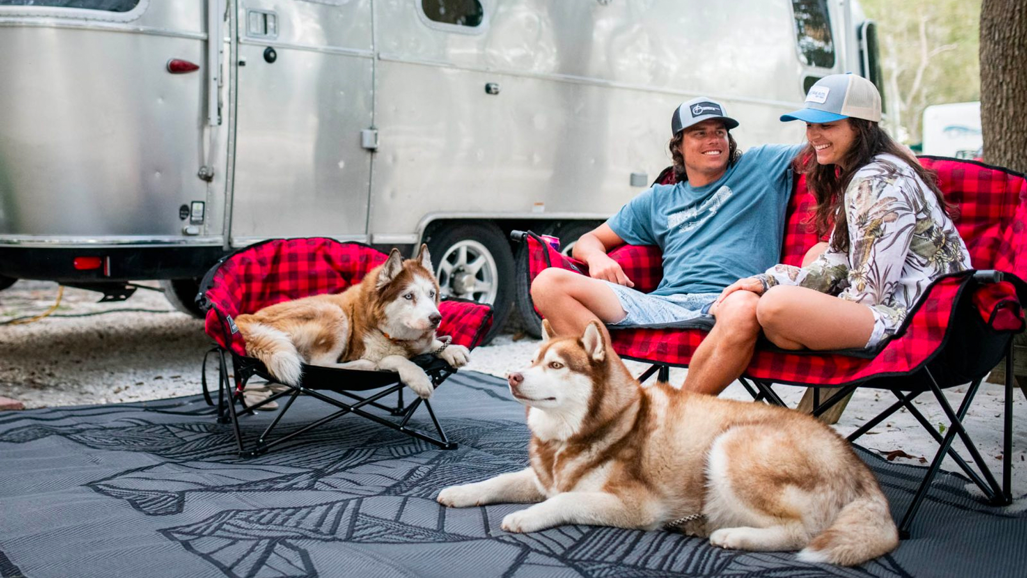 Two people sitting in a KUMA chair outside of an Airstream travel trailer with two dogs
