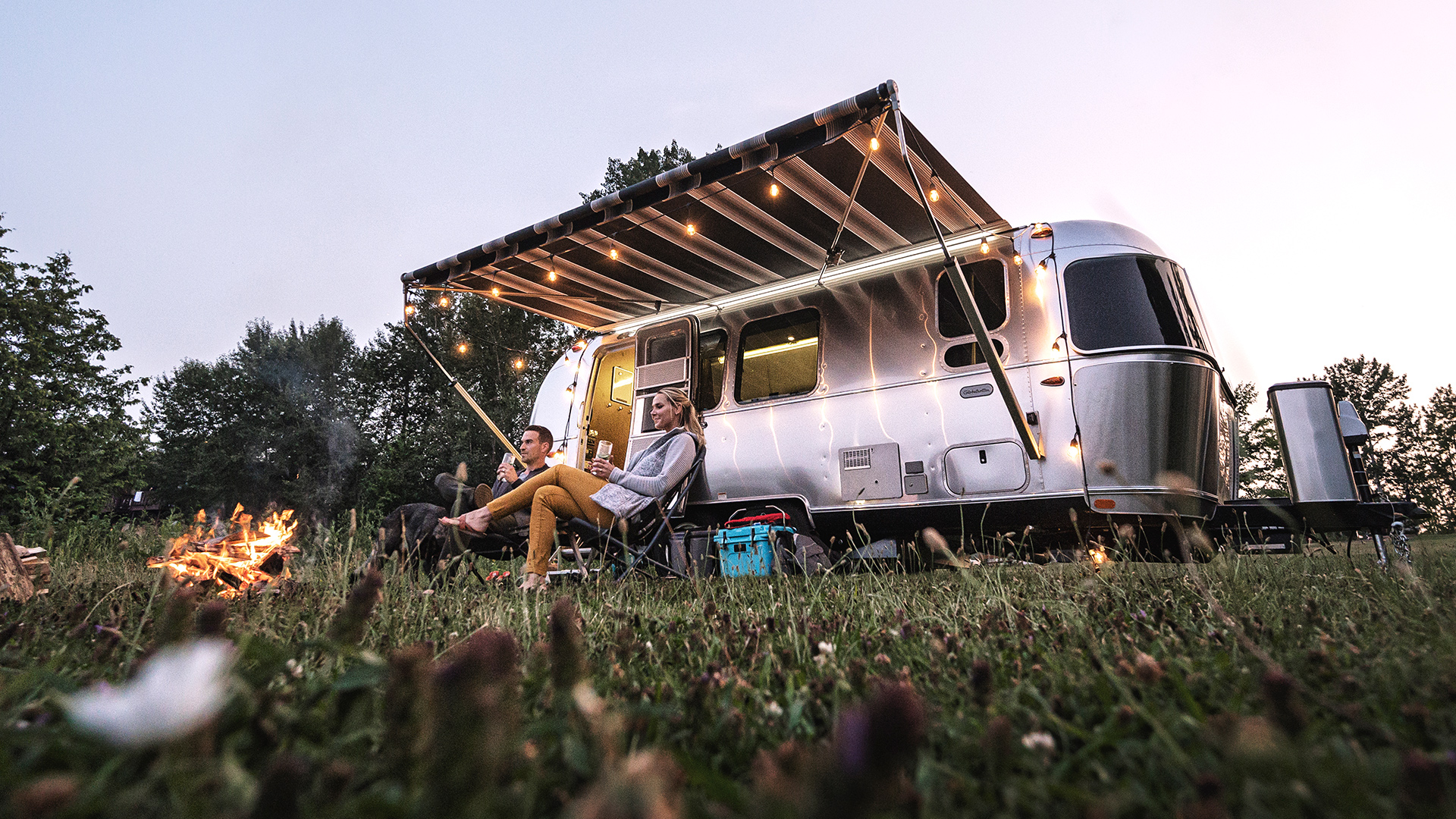 An Airstream Globetrotter travel trailer parked in a grass field with the awning out and people sitting outside.
