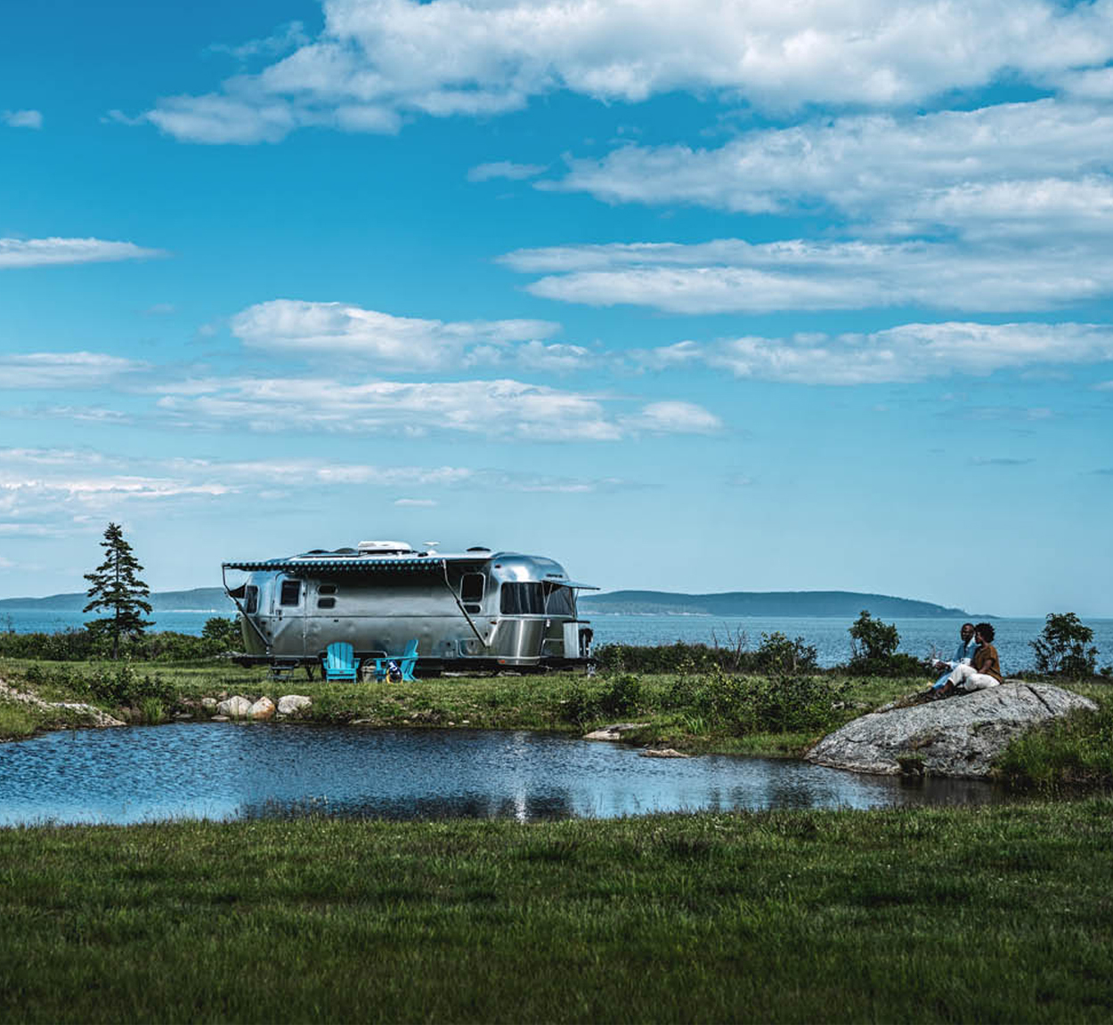 An Airstream International Travel Trailer parked by a pond with a couple sitting on a rock outside.