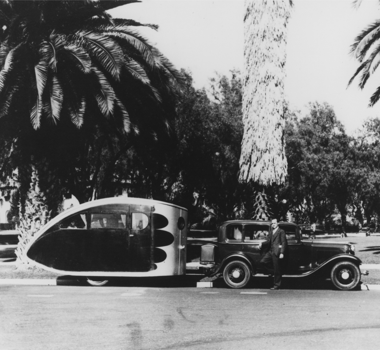 An Airstream Torpedo travel trailer being towed by a vintage car in a tropical area