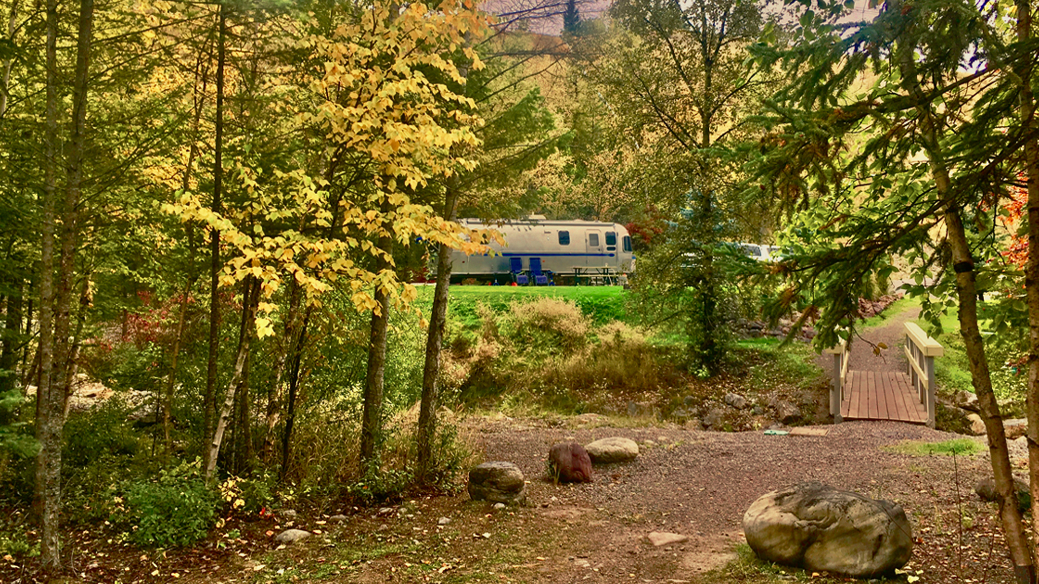 An Airstream Classic travel trailer parked in the woods with two chairs sitting outside.