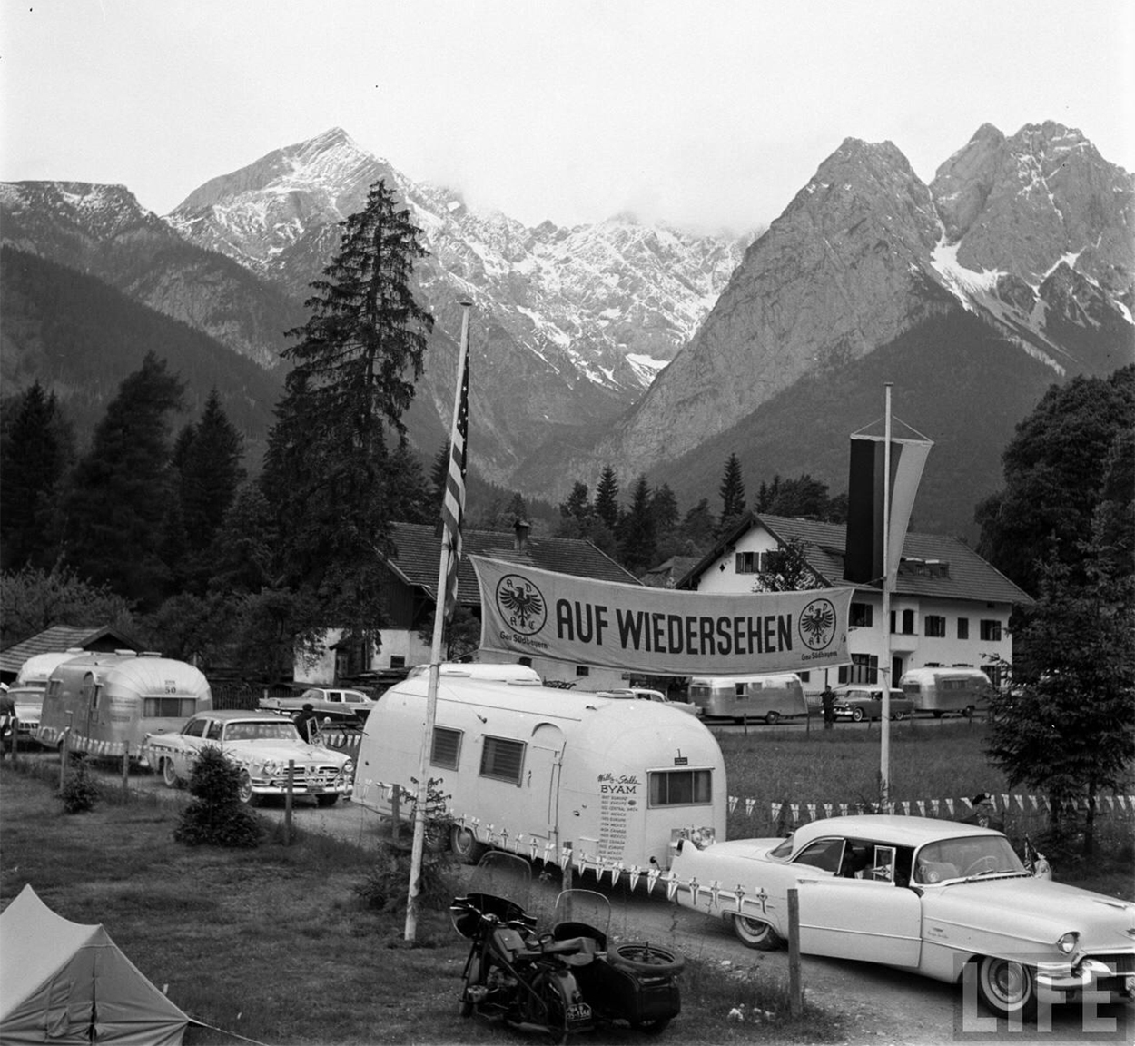 Vintage white trailer Airstream being towed in a white car by Airstream founder, Wally Byam and his wife Stella, as they travel through Europe.