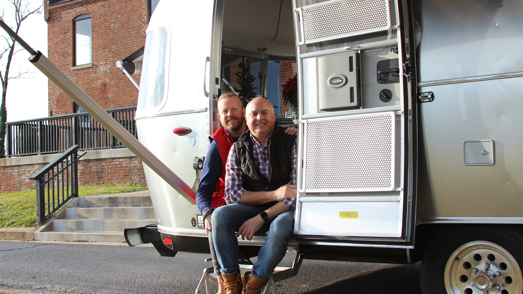 Bob Hoffman and Wayne sitting in the doorway of their Airstream travel trailer that is parked next to a brick building
