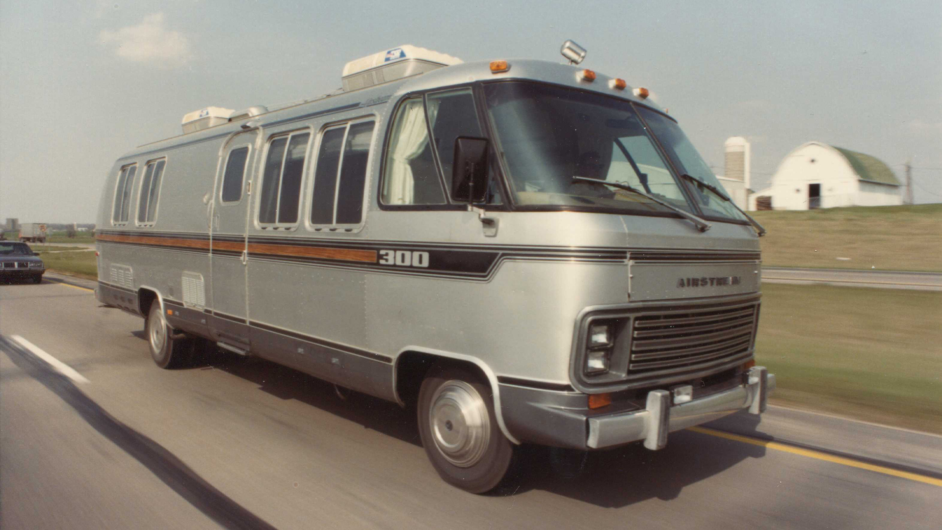 A vintage Airstream Classic Class B Motorhome driving down the road with a white barn and farm in the background.