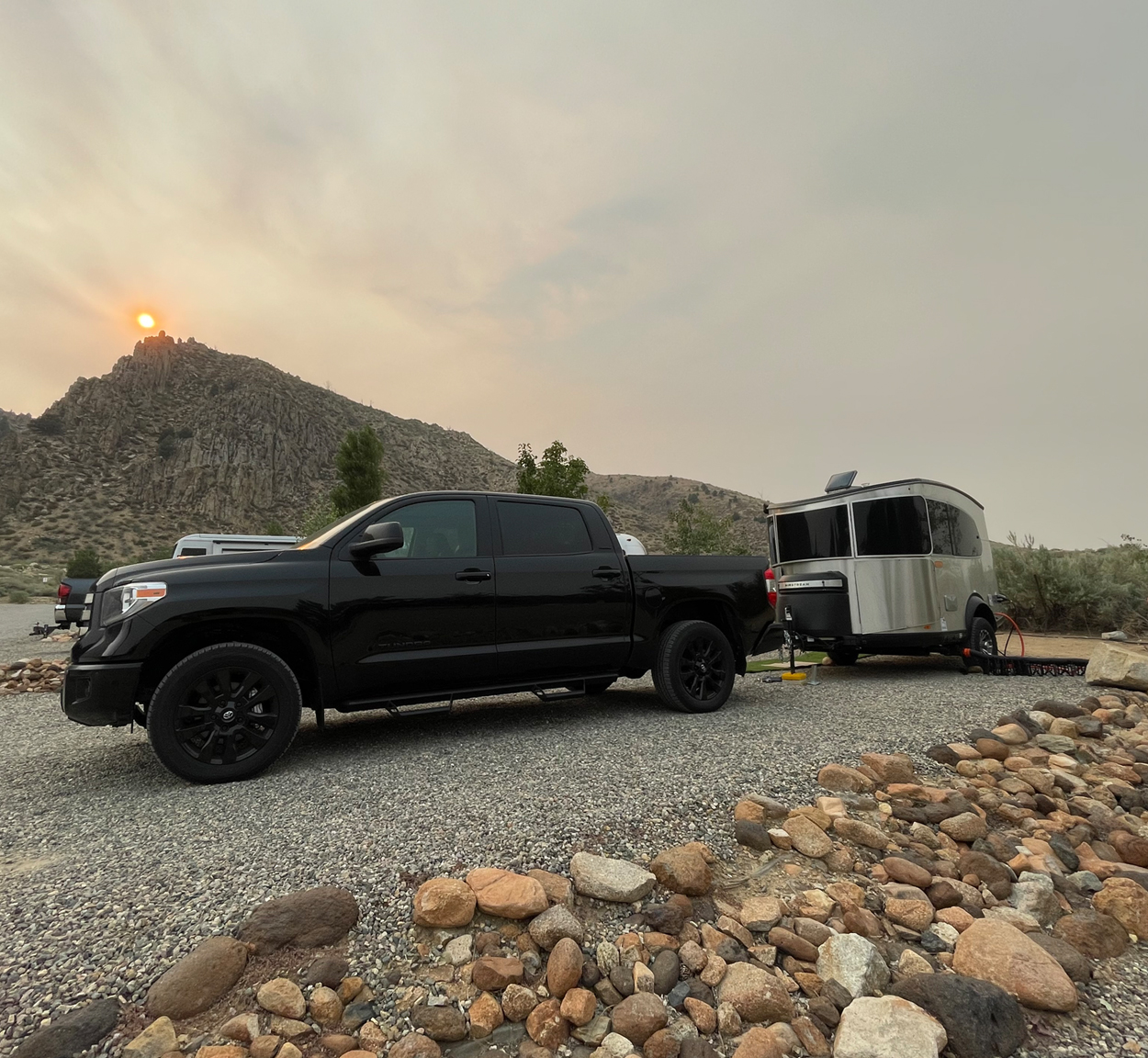 Black truck parking an Airstream Basecamp travel trailer at a campground with mountains and a sunset in the background