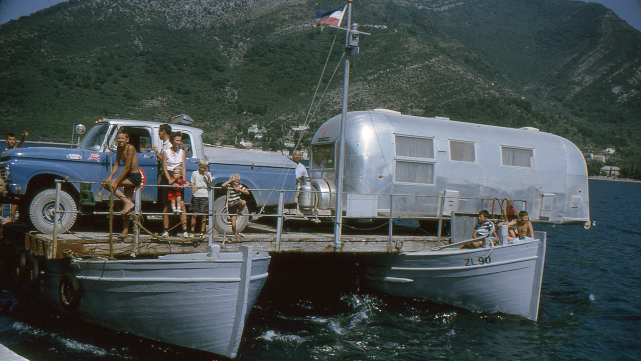 People on a boat that is carrying an Airstream travel trailer across the water during the Around the World Caravan.