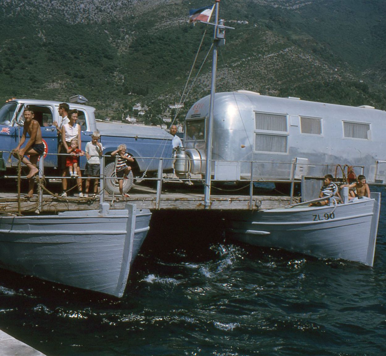 People on a boat that is carrying an Airstream travel trailer across the water during the Around the World Caravan.