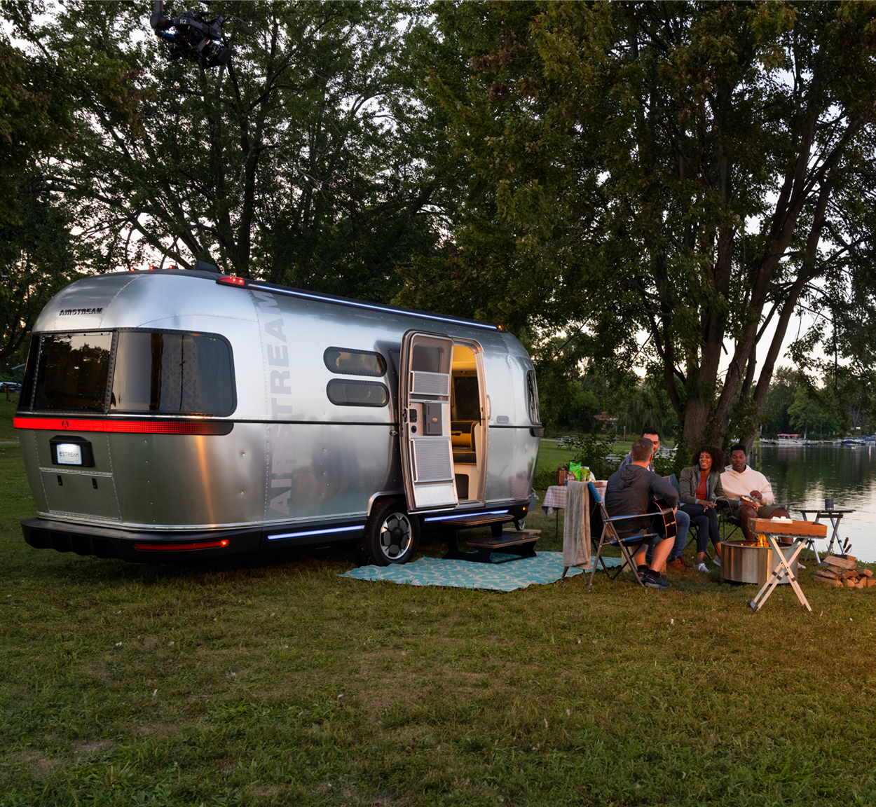 People sitting around a fire and by a lake and the Airstream all-electric Airstream travel trailer that is a concept vehicle.