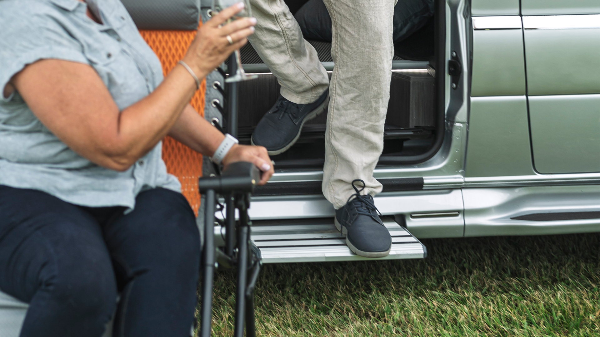 A person walking out of an Airstream Class B van while another person sits in a lawn chair outside of the touring coach.