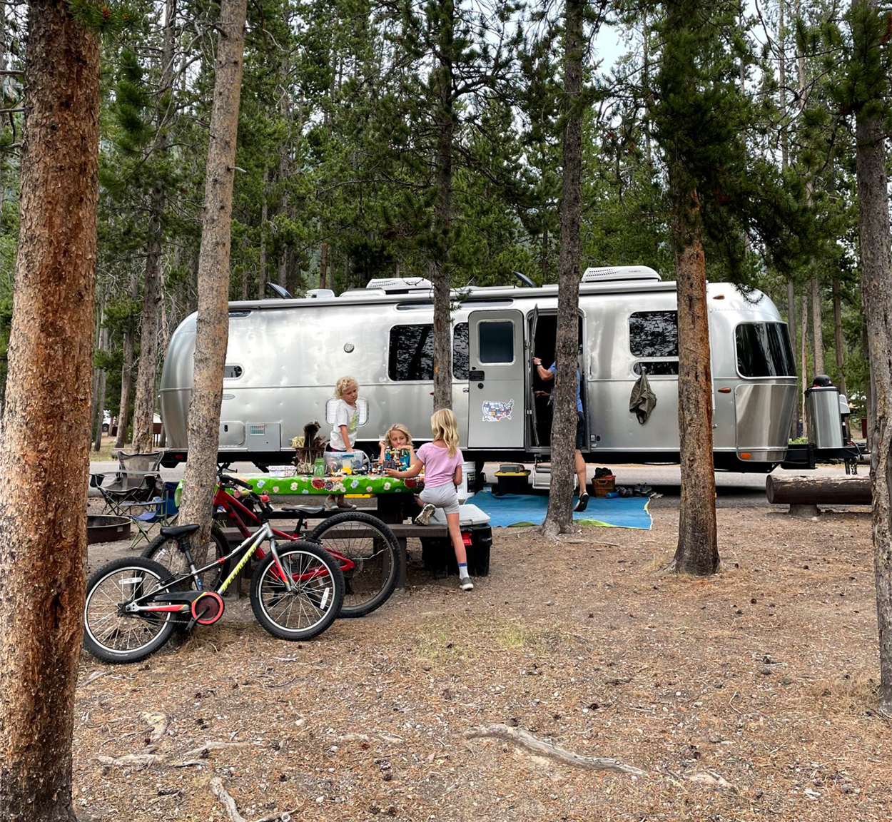 Kids sitting on a picnic table and playing outside of their Airstream travel trailer while they camp.