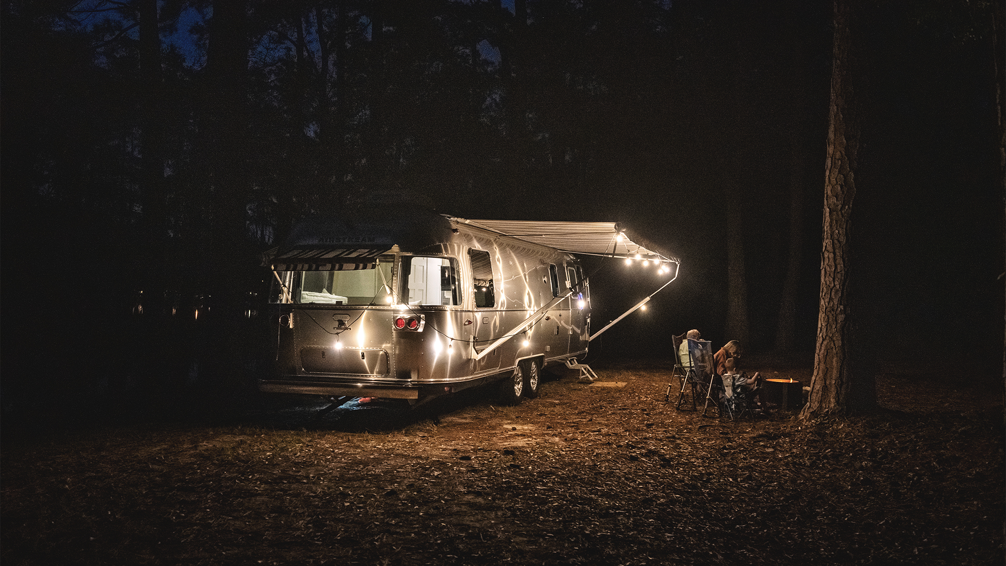 A family sitting outside by a campfire at night next to their Airstream Classic travel trailer while they are camping.