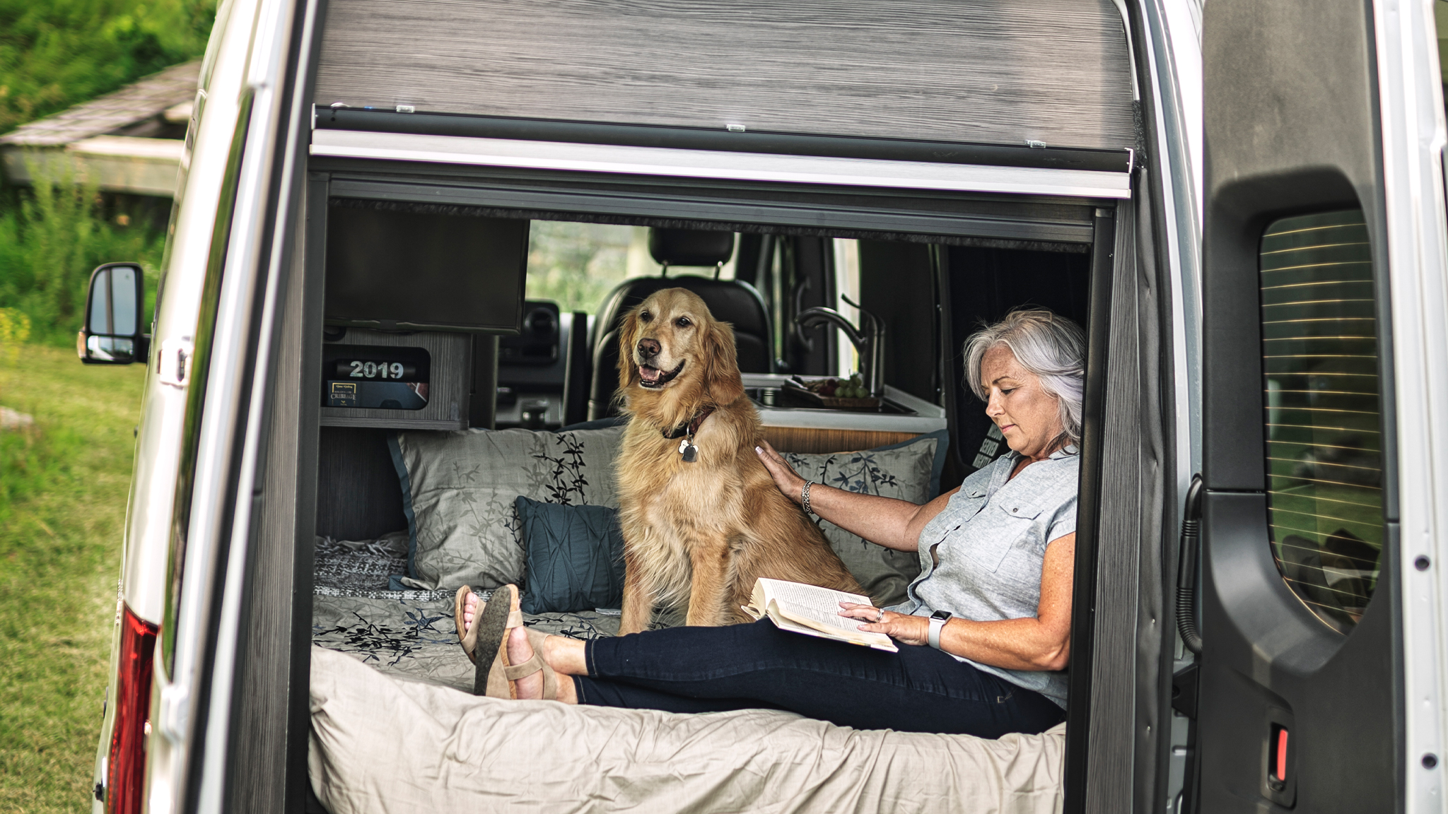 A woman and her dog sitting in the back of an Interstate 19 Class B motorhome while they sit in the bed while she reads with the rear doors open.
