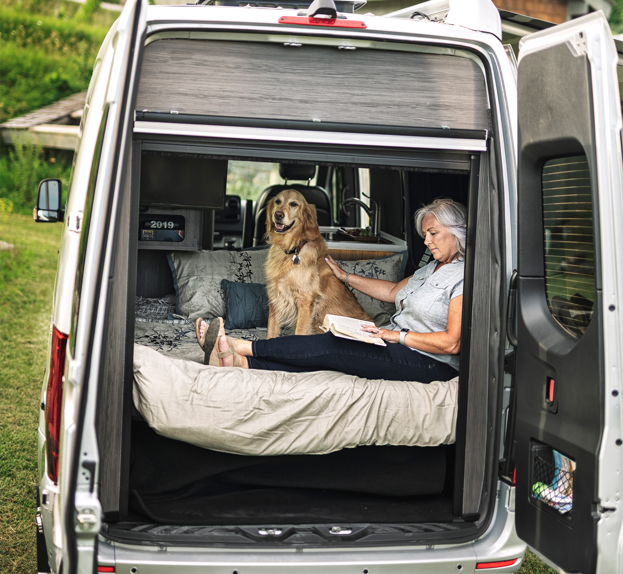 A woman and her dog sitting in the back of an Interstate 19 Class B motorhome while they sit in the bed while she reads with the rear doors open.