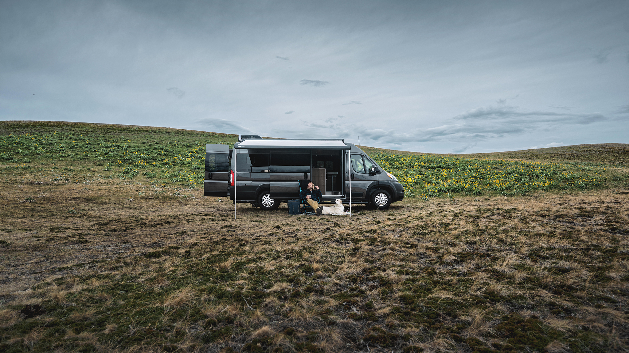 A man and his white dog sitting outside under the awning of their Airstream Rangeline Class B Motorhome that is parked in an open field.