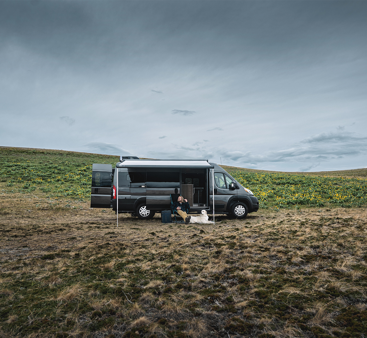 A man and his white dog sitting outside under the awning of their Airstream Rangeline Class B Motorhome that is parked in an open field.
