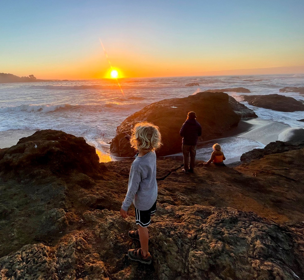 Kids and their parent standing on rocks by a body of water during a sunset.