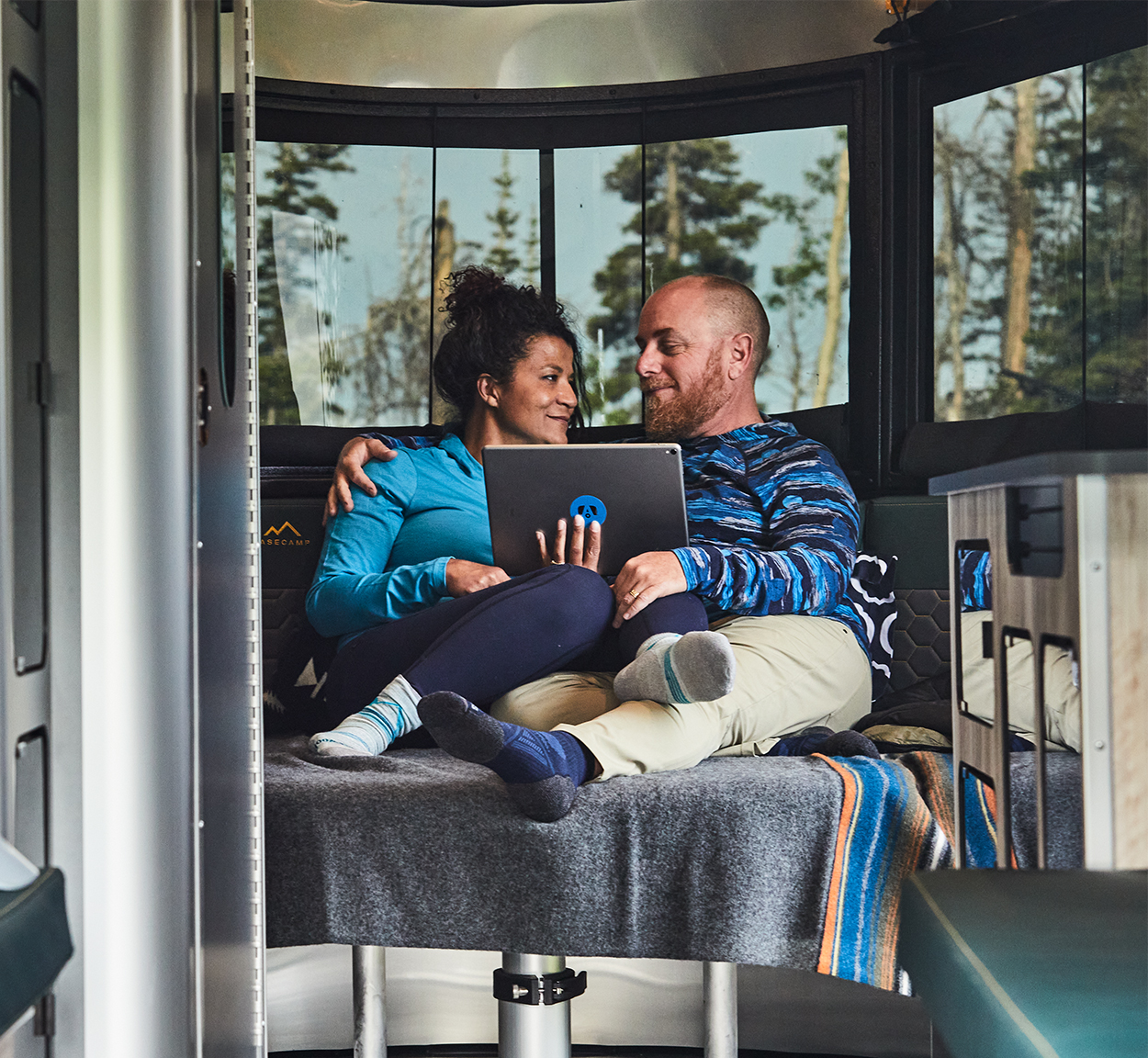 Two campers sitting on the bed of an Airstream Basecamp travel trailer watching their tablet.