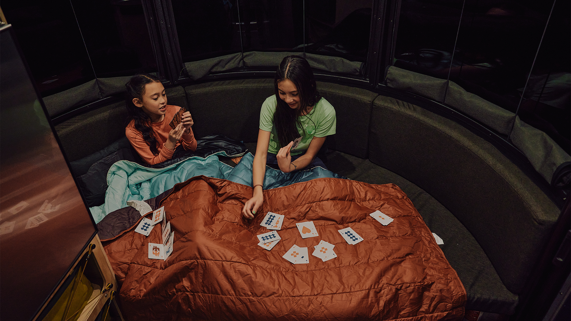 Two campers sitting on the bed at night playing cards in their Airstream REI Co-op Special Edition Basecamp 20X.